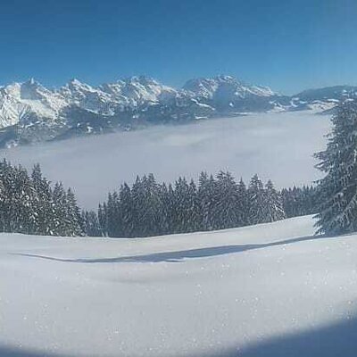 Die verschneite Berglandschaft rund um den Bauernhof mit Blick auf die Alpen und das Nebelmeer im Tal.
