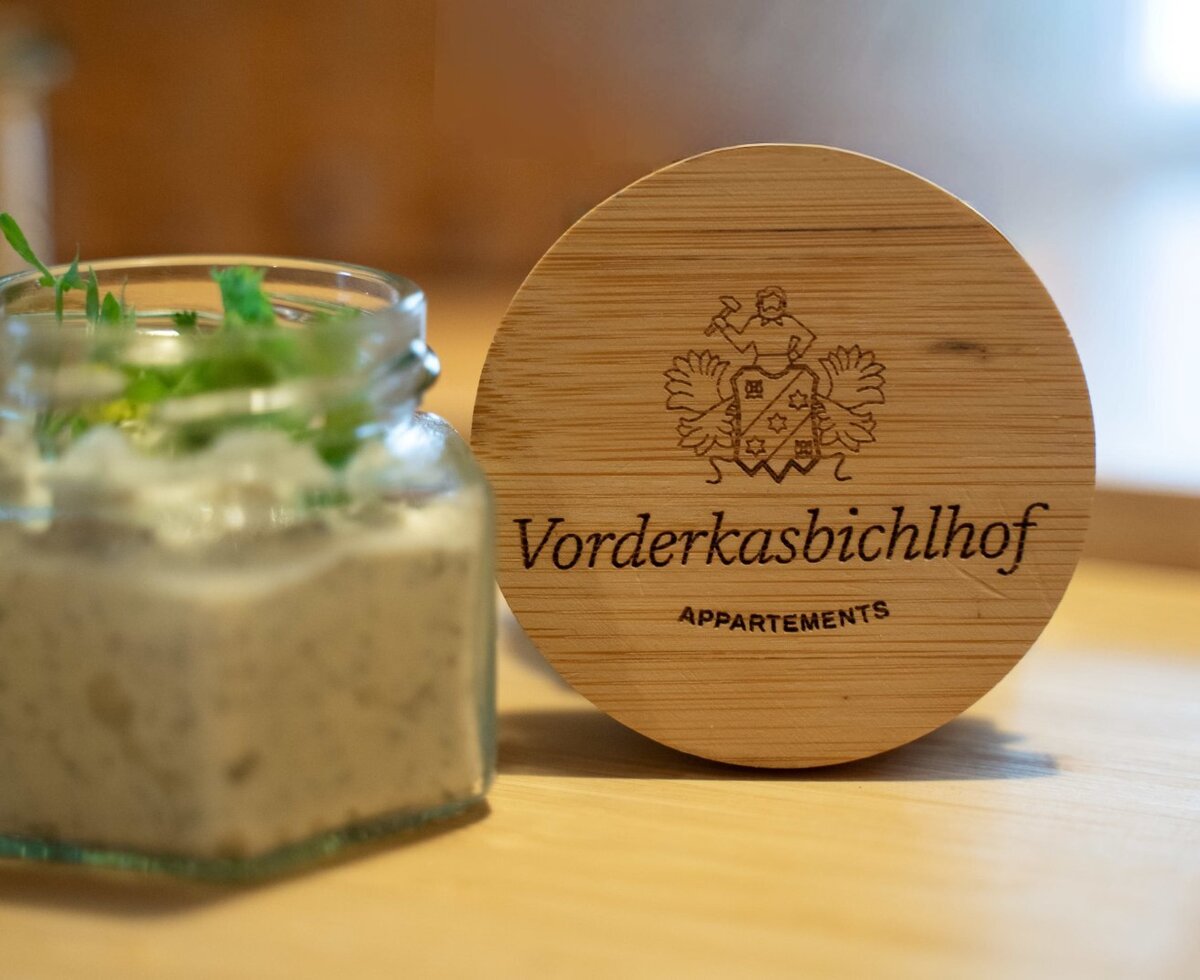 Ein Aufstrich im Glas, garniert mit Kräutern, steht neben einem Holzuntersetzer mit dem Logo des Bauernhofs.