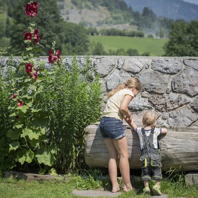 Ein Kind und eine Erwachsene im Außenbereich des Bauernhofs an einer Steinmauer mit Holzstamm, Blumen und Bergblick.