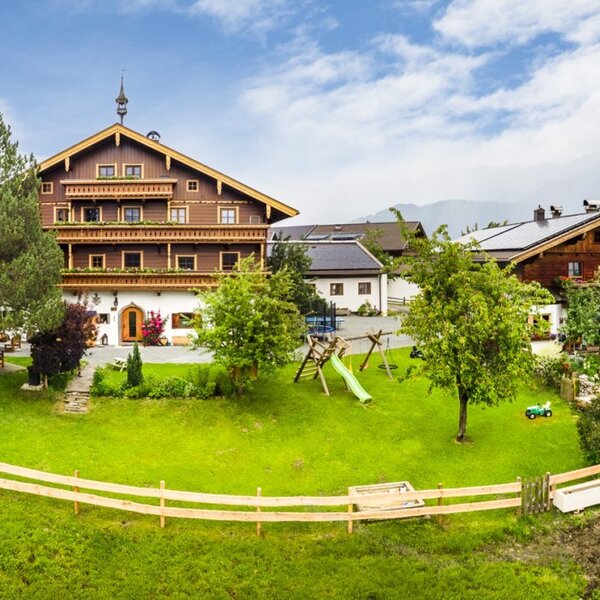 Ein einladender Bauernhof umgeben von einer weitläufigen grünen Wiese mit einem Kinderspielbereich und grasenden Kühen im Hintergrund.