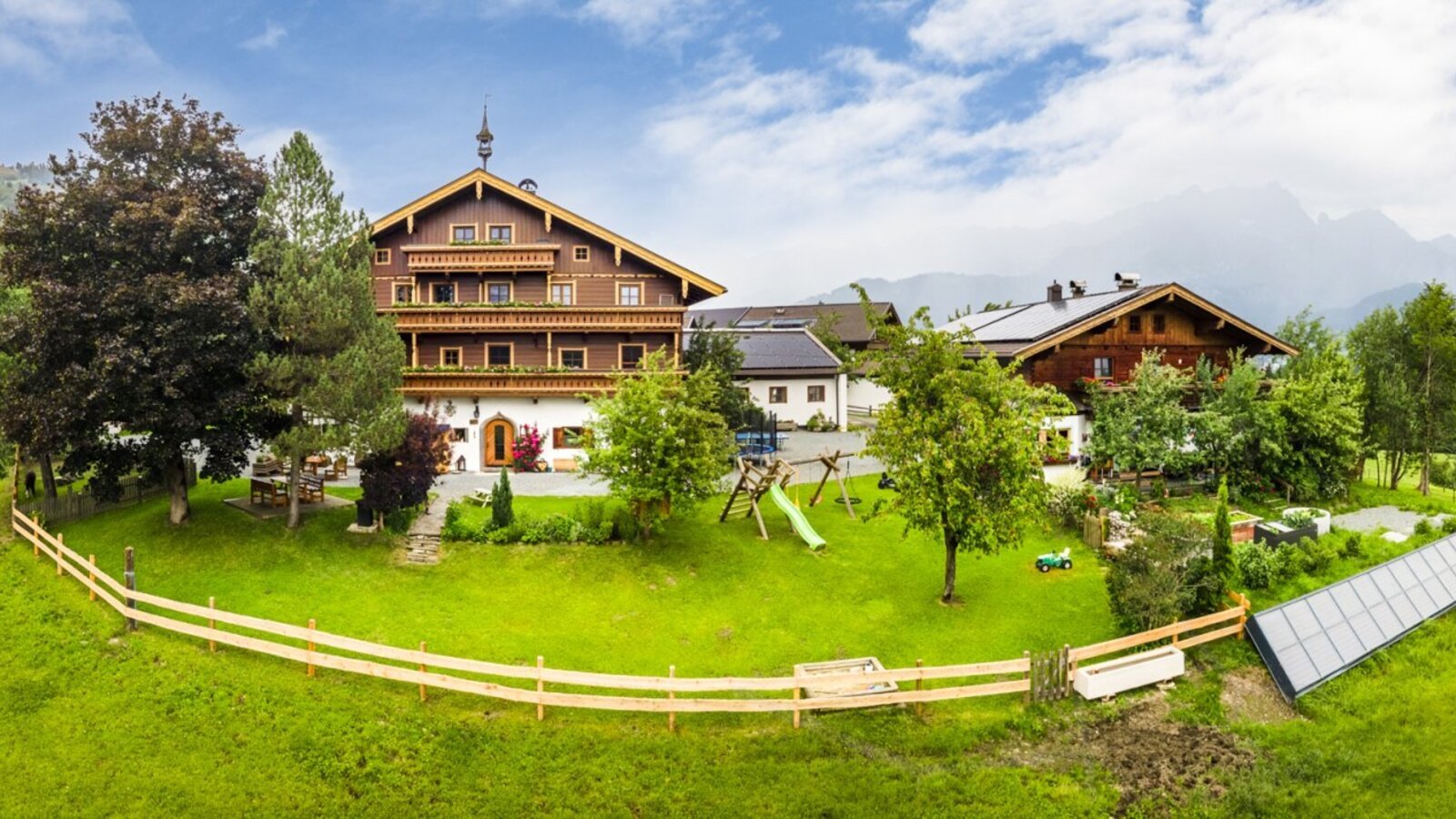 Ein einladender Bauernhof umgeben von einer weitläufigen grünen Wiese mit einem Kinderspielbereich und grasenden Kühen im Hintergrund.