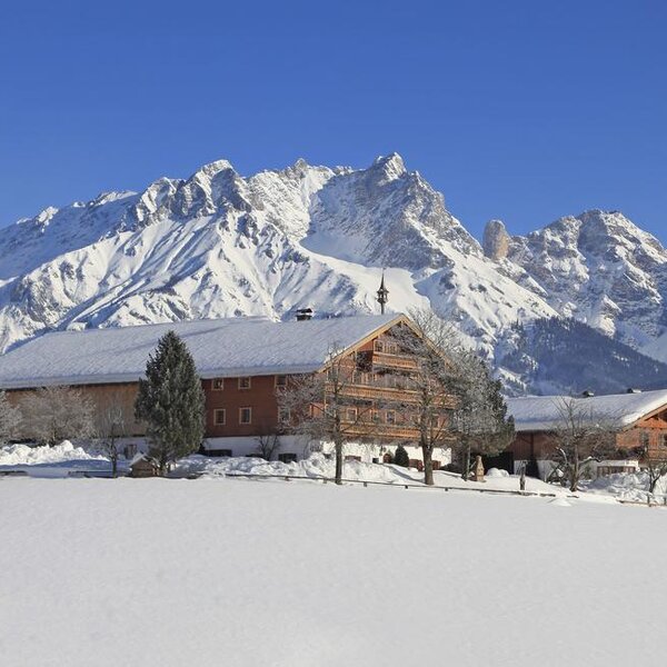 Der Bauernhof mit seinen traditionellen Holzbalkonen und schneebedeckten Dächern in einer Winterlandschaft vor einer Bergkulisse.
