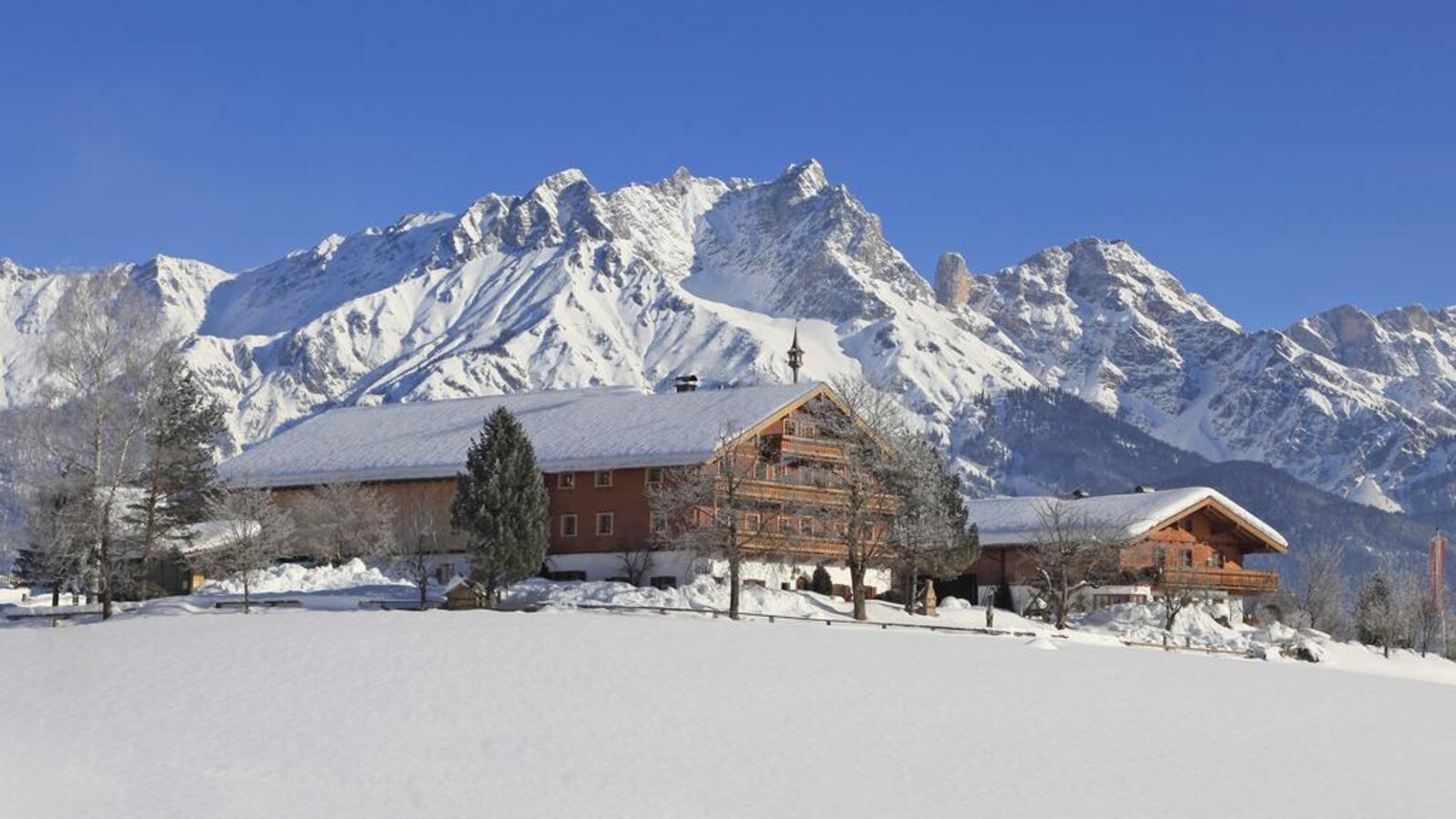 Der Bauernhof mit seinen traditionellen Holzbalkonen und schneebedeckten Dächern in einer Winterlandschaft vor einer Bergkulisse.
