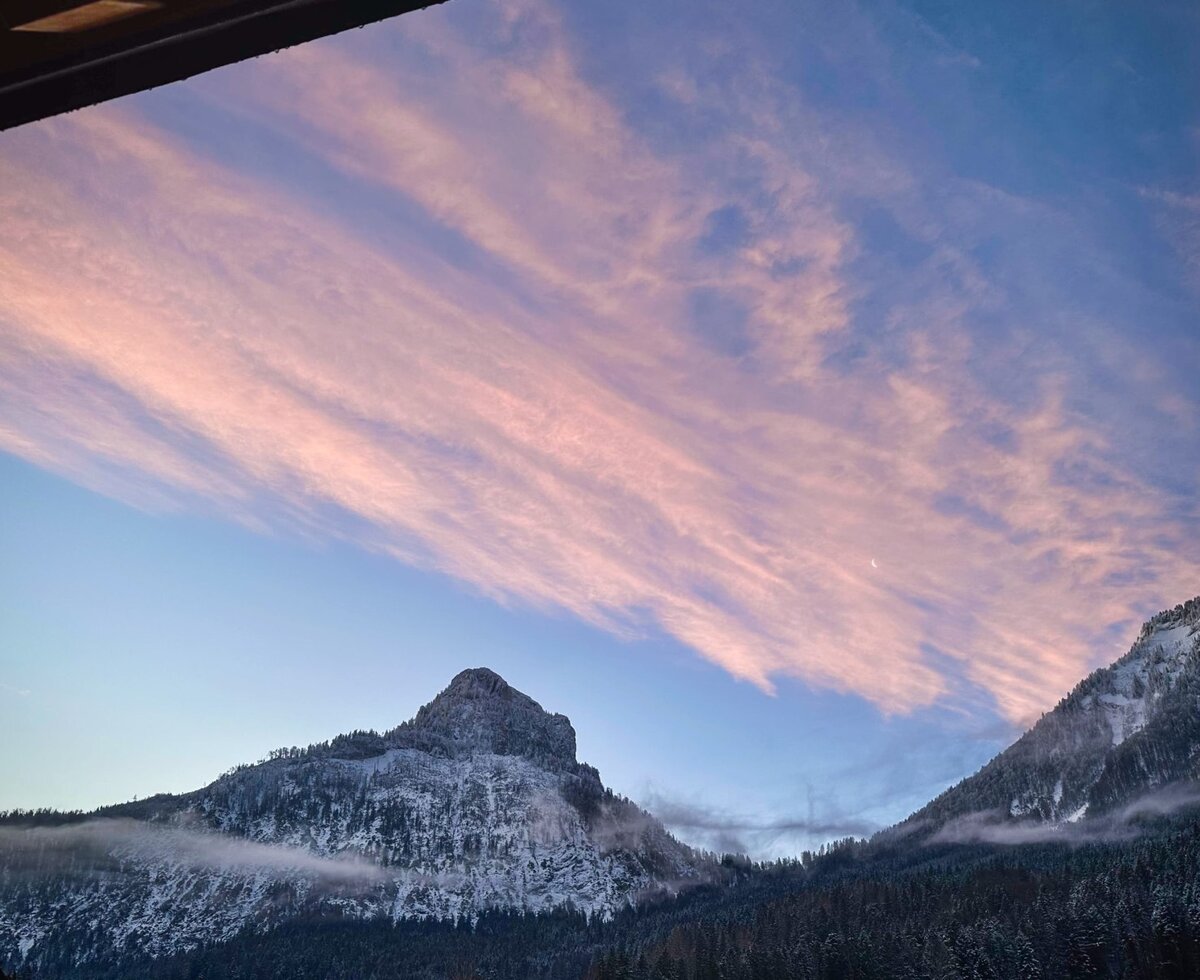 Der Blick vom Balkon des Bauernhofs auf schneebedeckte Berge unter einem Himmel mit rosa Wolken und einer Mondsichel.