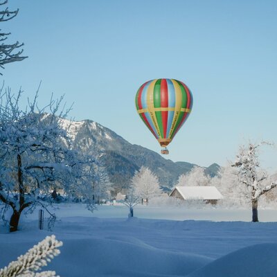 Winterliche Landschaft um den Bauernhof mit schneebedeckten Bäumen, Bergen und Heißluftballons am Himmel.