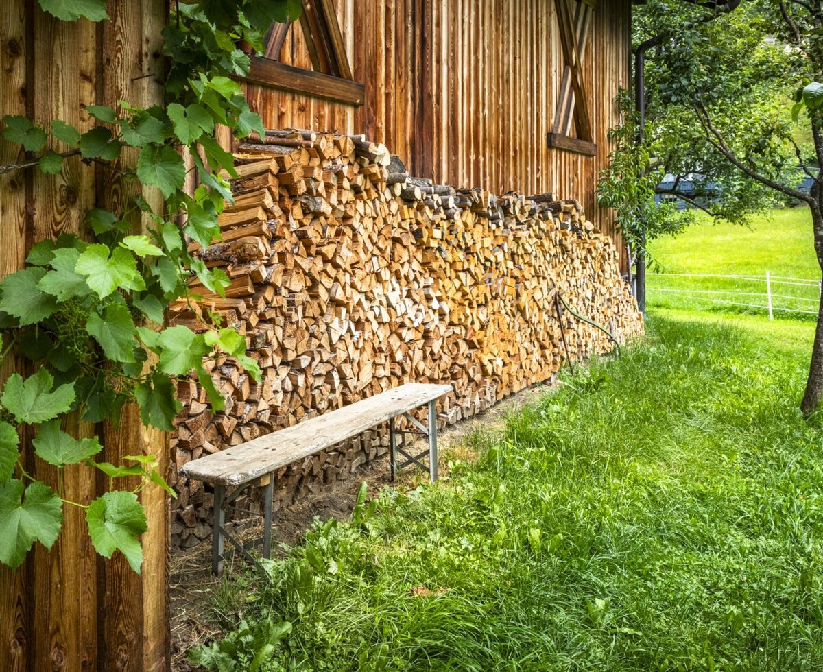 Ein idyllischer Außenbereich am Bauernhof mit einer urigen Holzbank vor ordentlich aufgeschichtetem Kaminholz und einer grünen Wiese.