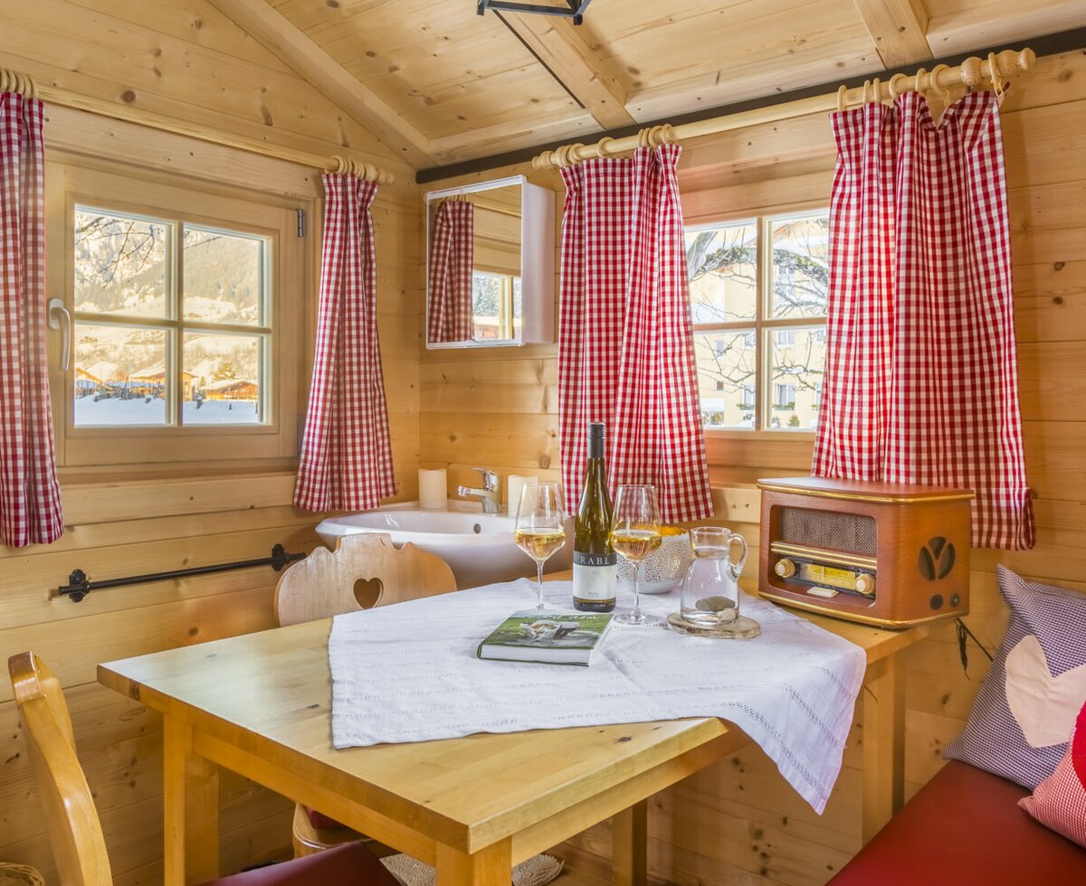 Essbereich in der Auszeithütte mit Holzwänden, einem Tisch für zwei Personen, Spülbecken, Fenstern mit karierten Vorhängen und einem Radio.