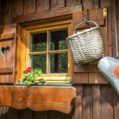 Holzfenster der Auszeithütte mit Blumenkasten, roten Geranien und Fensterläden mit Herzmotiv.
