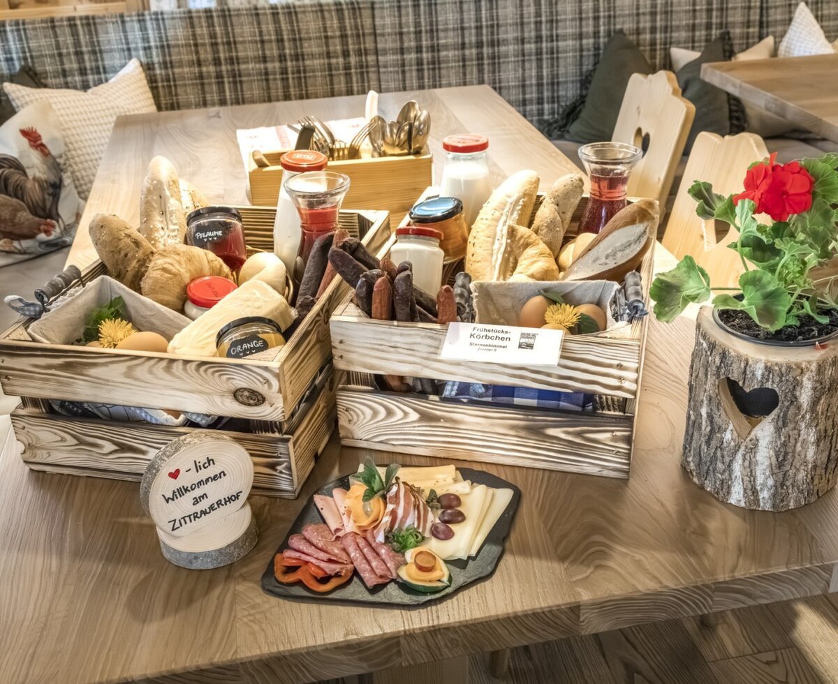 Frühstück auf dem Tisch, serviert in Holzkisten mit Brot, Gebäck, Aufstrichen, Getränken und Eiern, ergänzt durch eine Platte mit Wurst und Käse für die Gäste der Ap­par­te­ments.