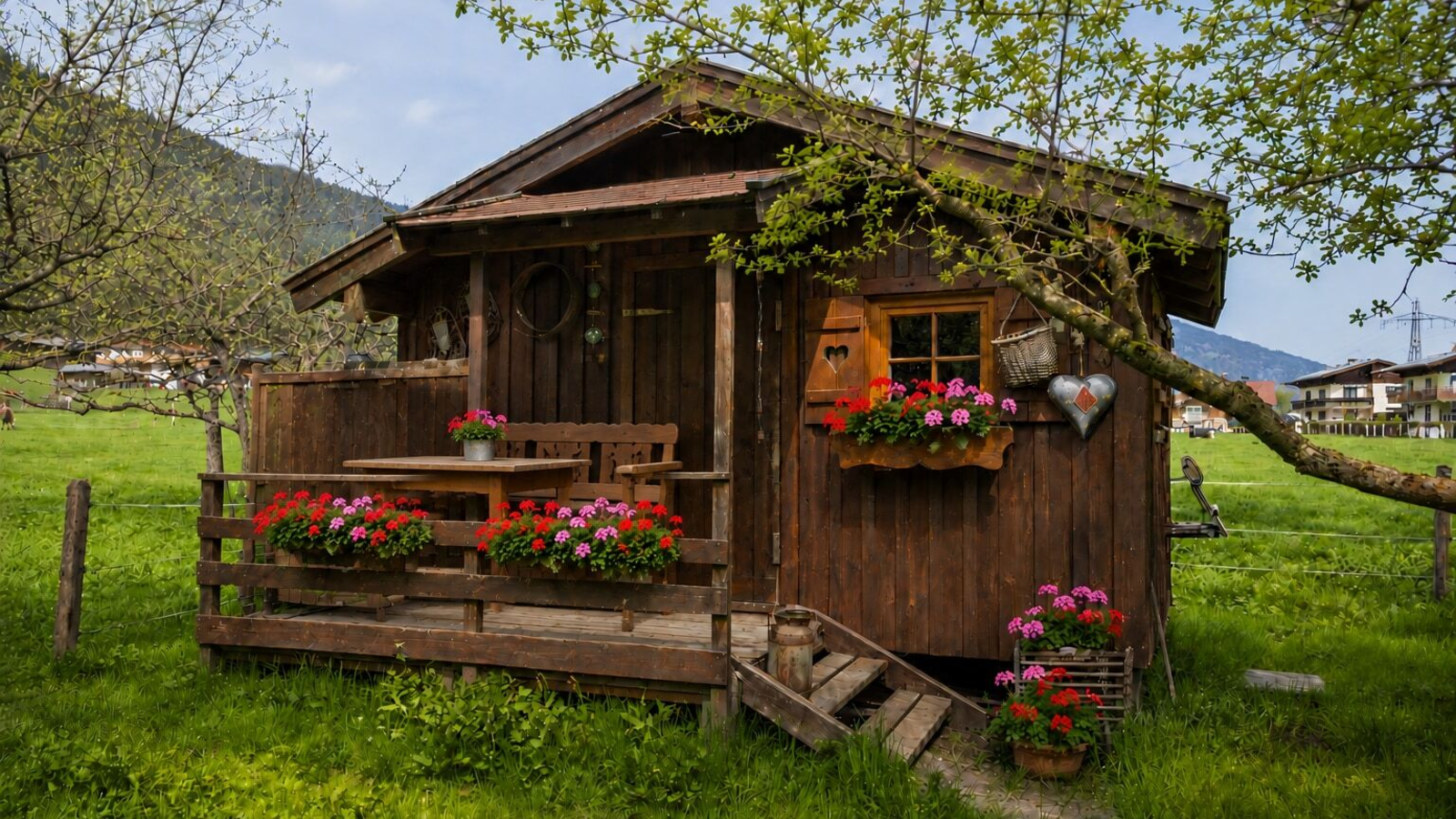 Gartenhütte aus Holz mit einer kleinen Terrasse, ausgestattet mit Holztisch und Bänken, umgeben von einer grünen Wiese und Blumenkästen.