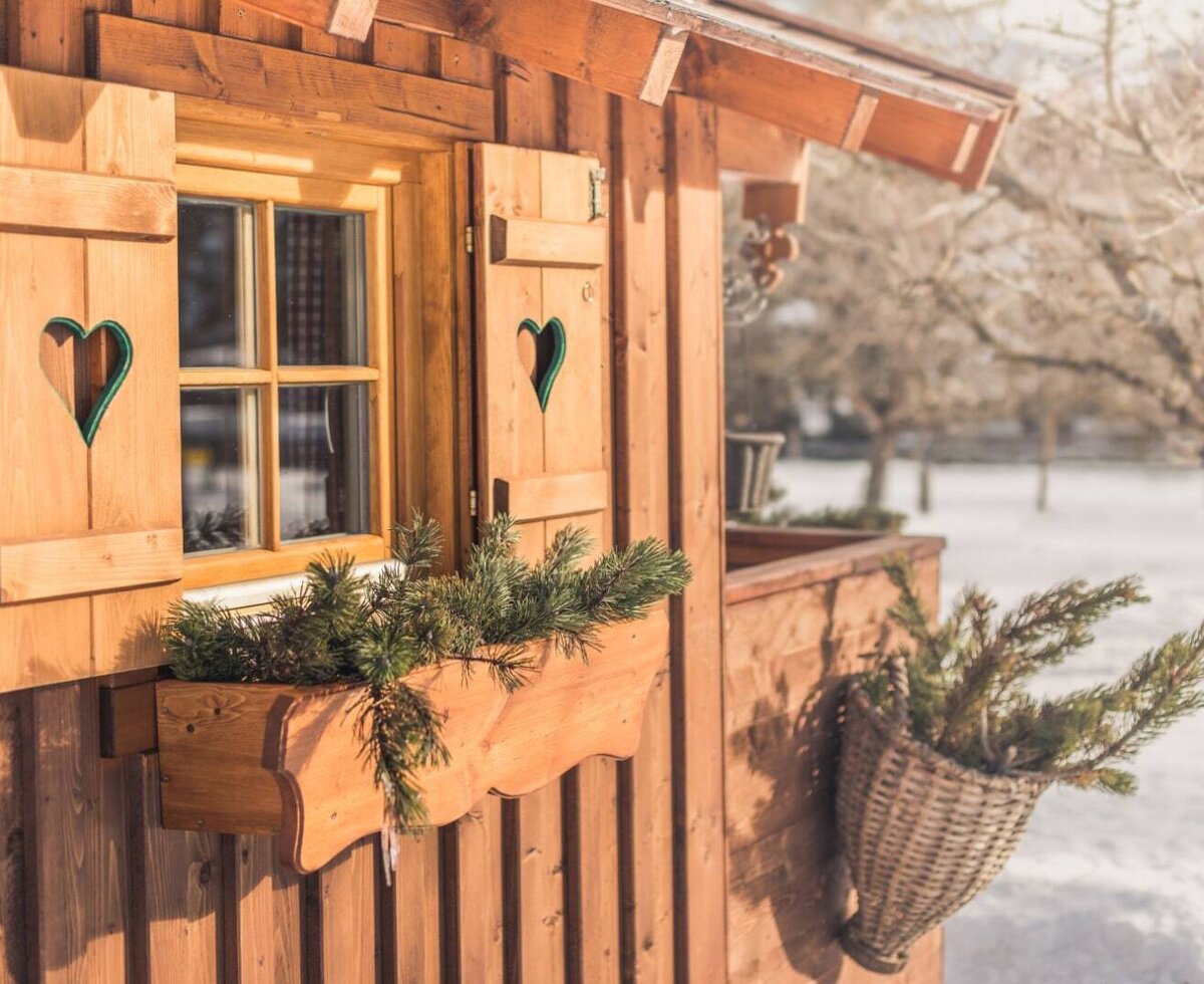 Ein Fenster des Ap­par­te­ments mit Holzfensterläden, Herzmotiv und einem Blumenkasten mit Tannenzweigen.