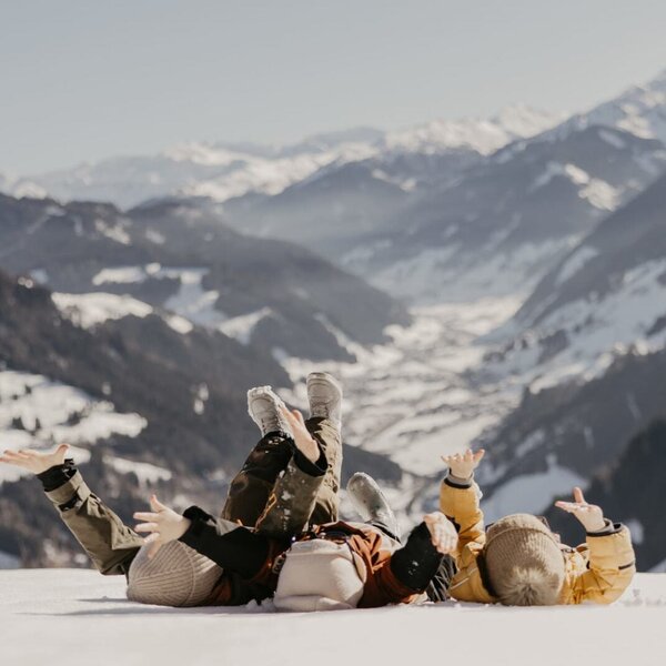 Fröhliche Kinder beim Spielen im Schnee nahe dem Bauernhof mit einer beeindruckenden, verschneiten Bergkulisse im Hintergrund.