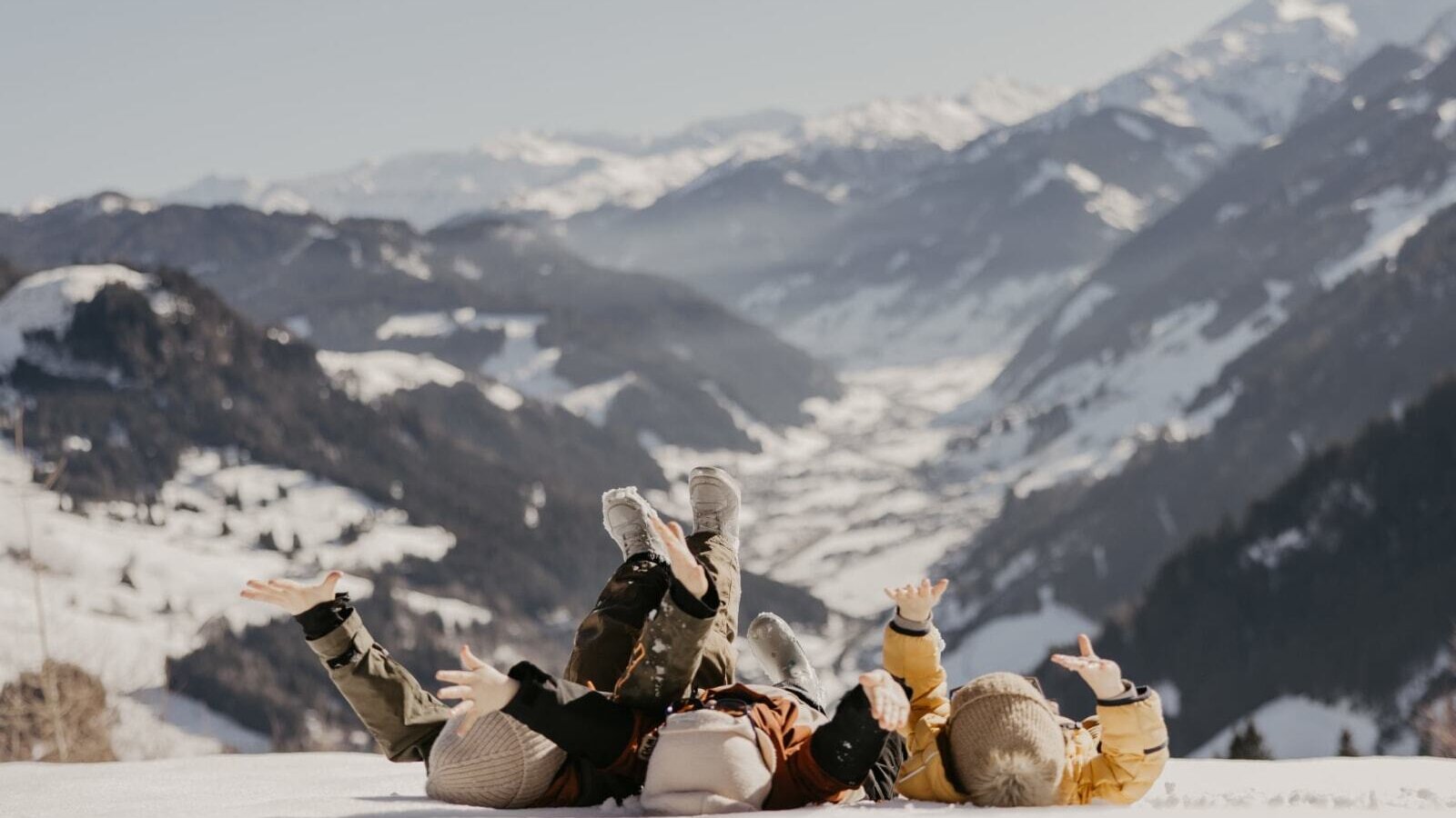 Fröhliche Kinder beim Spielen im Schnee nahe dem Bauernhof mit einer beeindruckenden, verschneiten Bergkulisse im Hintergrund.