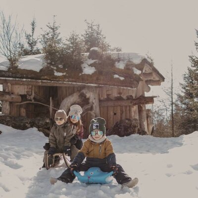 Ein winterliches Rodelabenteuer für Kinder im Schnee direkt am Bauernhof mit einer urigen Holzhütte im Hintergrund.