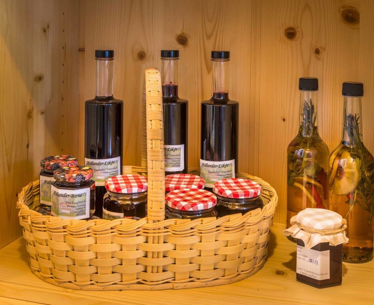 Ein einladender Weidenkorb im Holzschrank des Bauernhofs, gefüllt mit hausgemachten Likören und Marmeladen mit rot-weiß karierten Deckeln.