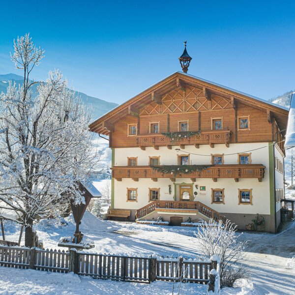 Der Bauernhof in winterlicher Umgebung mit traditioneller Holzfassade, Balkonen und Blick auf die verschneiten Alpen.