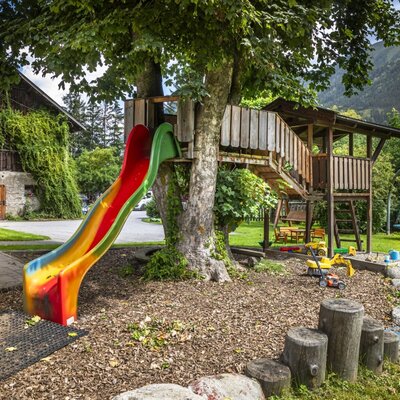 Der einladende Kinderspielplatz des Bauernhofs bietet eine bunte Rutsche, ein Baumhaus und eine Schaukel vor einer malerischen Bergkulisse.