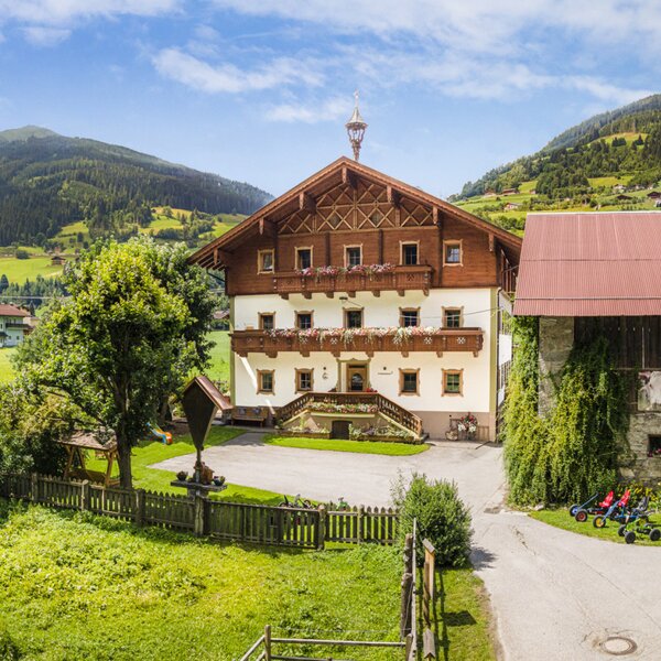 Der einladende Bauernhof im traditionellen Alpendesign, umgeben von einer weitläufigen grünen Wiese und den sanften Berghängen.