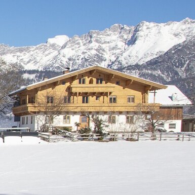 Der Bauernhof mit Holzfassade, umgeben von Schnee, vor der Kulisse schneebedeckter Berge.