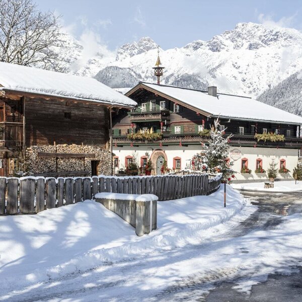 Außenansicht des Bauernhofs im Winter mit verschneiten Dächern, traditioneller Holzbauweise und Bergen im Hintergrund.