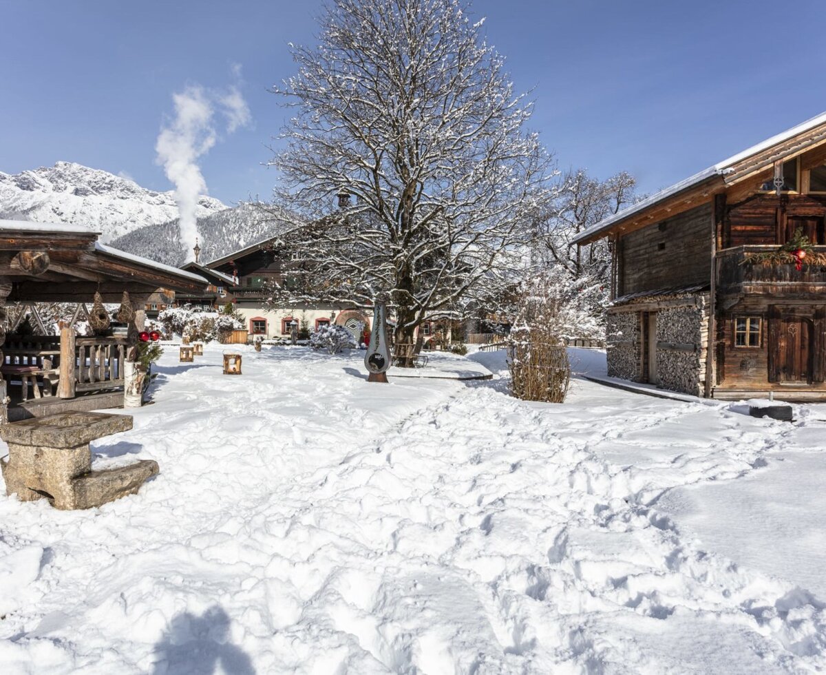 Der verschneite Außenbereich des Bauernhofs mit traditionellen Gebäuden, einem großen Baum und Bergen im Hintergrund.