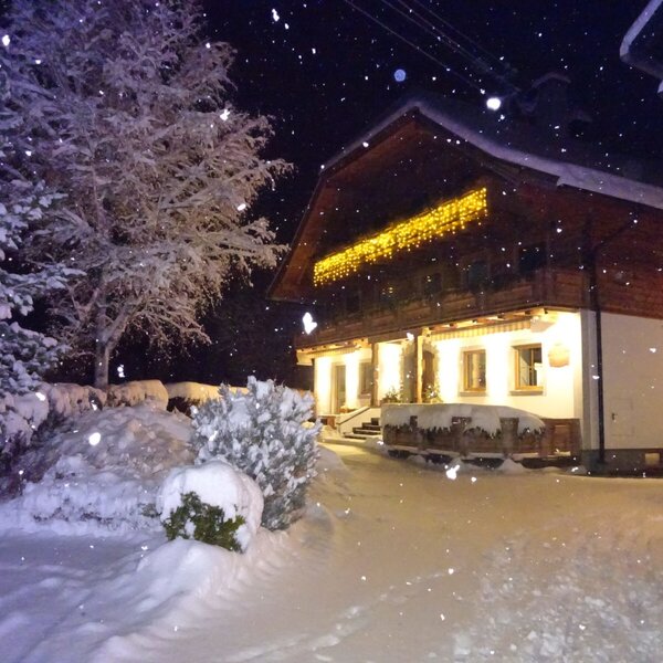 Der schneebedeckte Bauernhof bei Nacht mit fallendem Schnee und beleuchtetem Balkon.