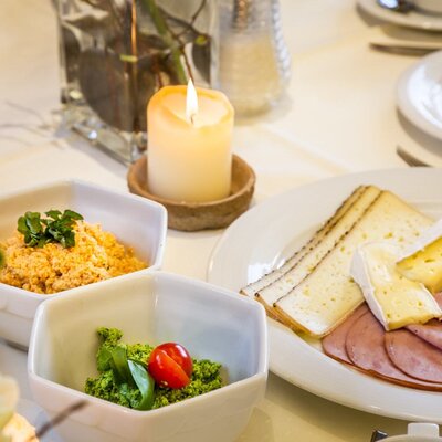 Ein gedeckter Frühstückstisch auf dem Bauernhof mit Käse, Wurst, zwei Aufstrichen und einer brennenden Kerze.