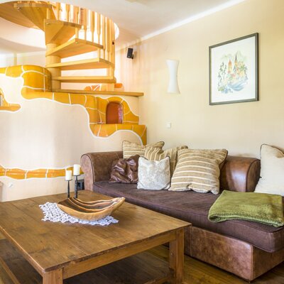 Der Wohnbereich des Bauernhofs mit Sofa, Holztisch, Kachelofen und Holz-Wendeltreppe.