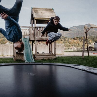 Kinder springen auf dem Trampolin des Bauernhofs, mit einem Spielplatz und Schaukeln im Hintergrund.