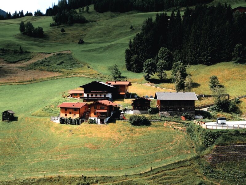 Der Bauernhof mit mehreren Gebäuden, umgeben von grünen Hügeln und Wäldern, inklusive Parkplatz.
