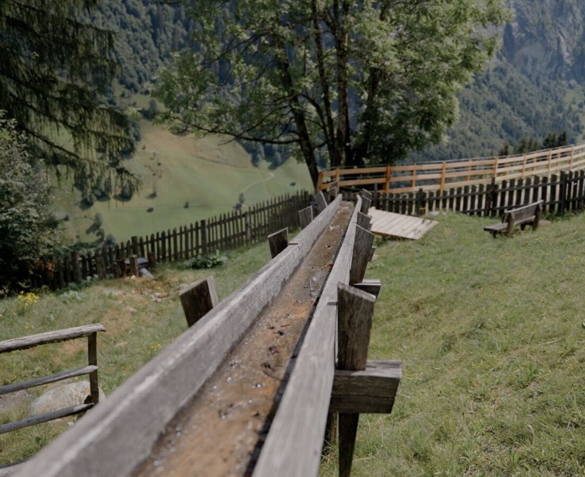 Hölzerne Wasserrinne auf dem Bauernhof mit Bergblick und einer grünen Wiese.