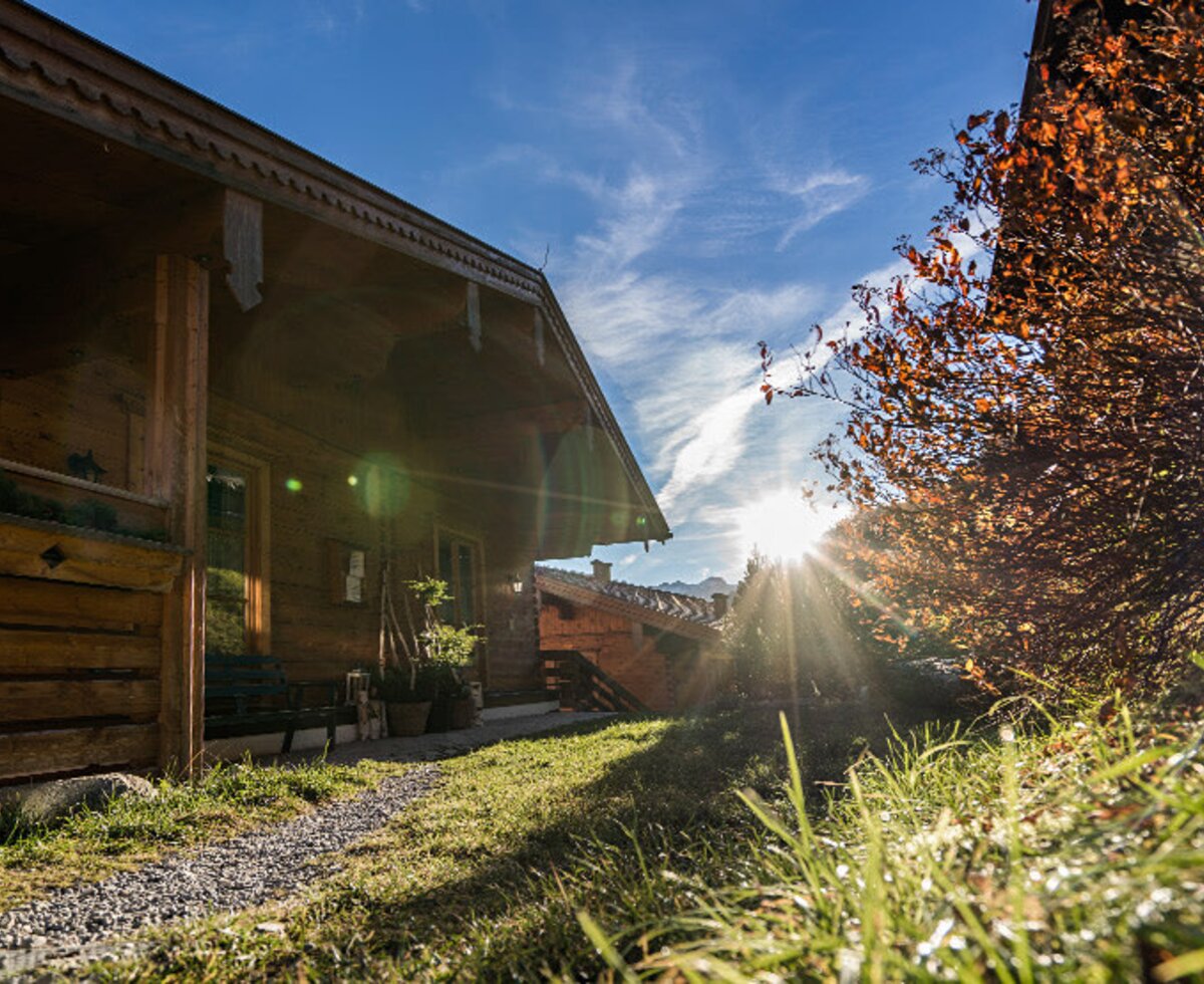 Die Holzfassade des Bauernhofs mit umgebender Wiese und Herbstlaub, im Sonnenlicht.