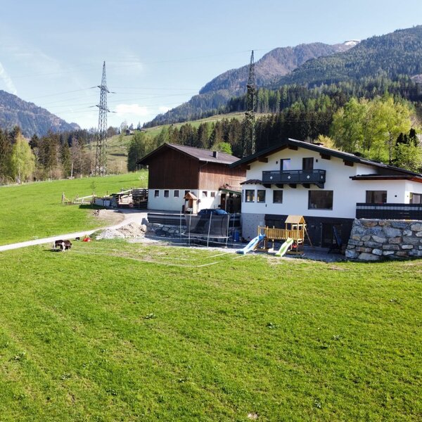 Außenansicht des Bauernhofs mit umgebenden grünen Wiesen, Bergkulisse und einem Spielplatz mit Trampolin.