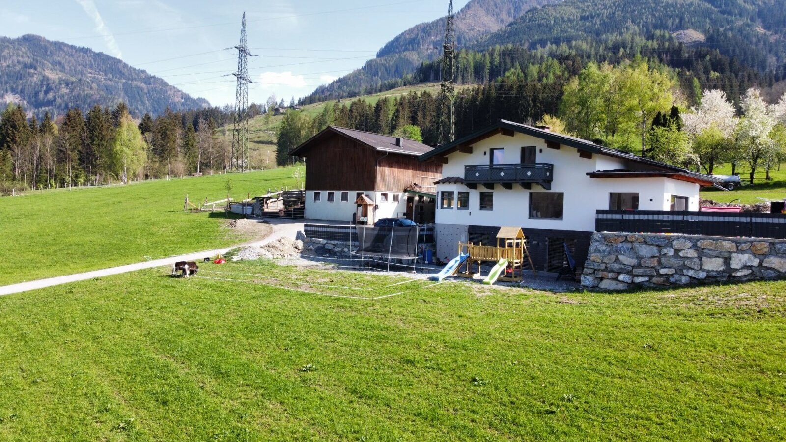 Außenansicht des Bauernhofs mit umgebenden grünen Wiesen, Bergkulisse und einem Spielplatz mit Trampolin.