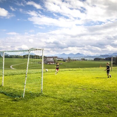Der Fußballplatz auf dem Bauernhof mit zwei Toren und spielenden Kindern vor der Bergkulisse.