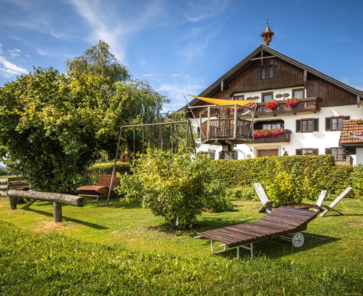 Der einladende Garten des Bauernhofs mit einer großen Liegewiese, Sonnenliegen und einem Kinderspielplatz vor dem traditionellen Haupthaus.