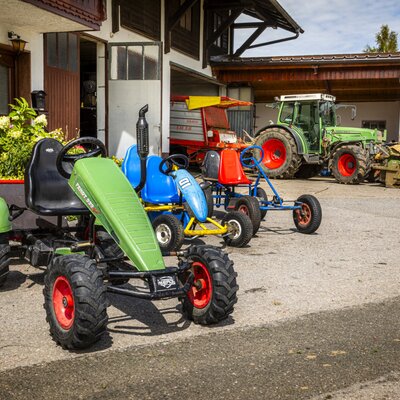Der einladende Außenbereich des Bauernhofs mit bunten Tretautos für Kinder und landwirtschaftlichen Fahrzeugen im Hintergrund.