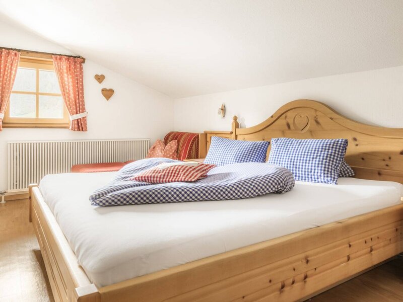 Schlafzimmer im Bauernhof mit Doppelbett, karierter Bettwäsche, Holzmöbeln und einer Sitzgelegenheit unter dem Fenster.