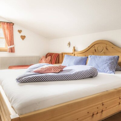Schlafzimmer im Bauernhof mit Doppelbett, karierter Bettwäsche, Holzmöbeln und einer Sitzgelegenheit unter dem Fenster.