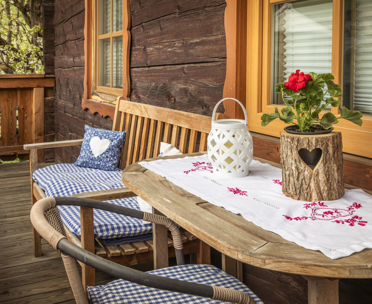 Der Balkon des Bauernhofs mit Holztisch, Sitzbank, karierten Kissen und einer roten Geranie.