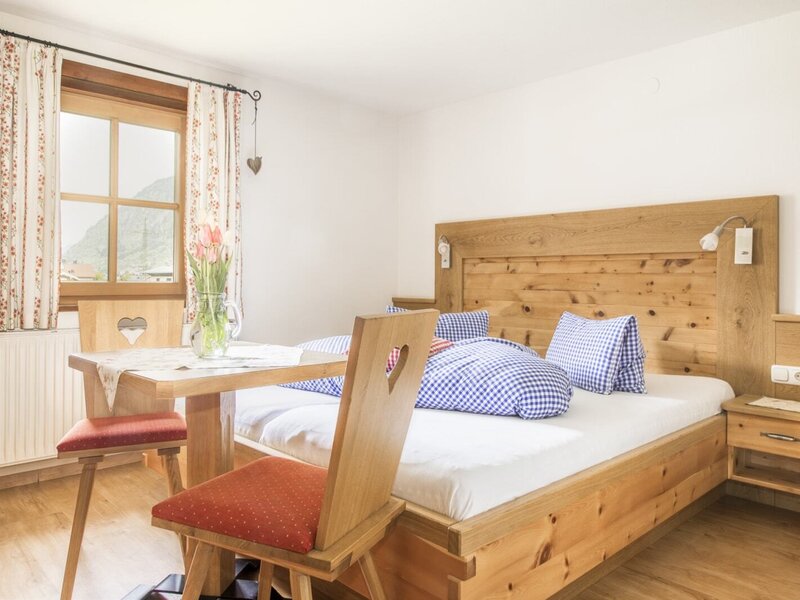 Doppelbettzimmer im Bauernhof mit Holzbett, Nachttisch, Tisch und Stühlen sowie einem Fenster mit Bergblick.