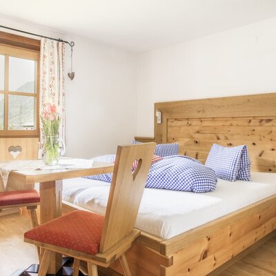 Doppelbettzimmer im Bauernhof mit Holzbett, Nachttisch, Tisch und Stühlen sowie einem Fenster mit Bergblick.