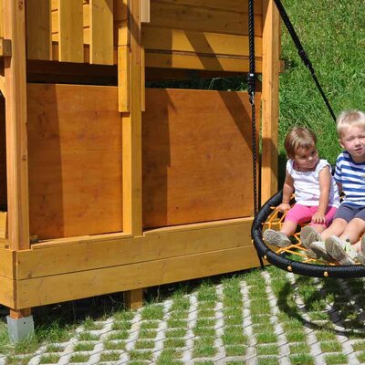 Kinder auf der Nestschaukel am Spielplatz der Ferienwohnung mit Holzspielhaus.