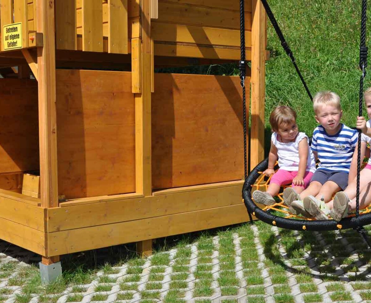 Kinder auf der Nestschaukel am Spielplatz der Ferienwohnung mit Holzspielhaus.