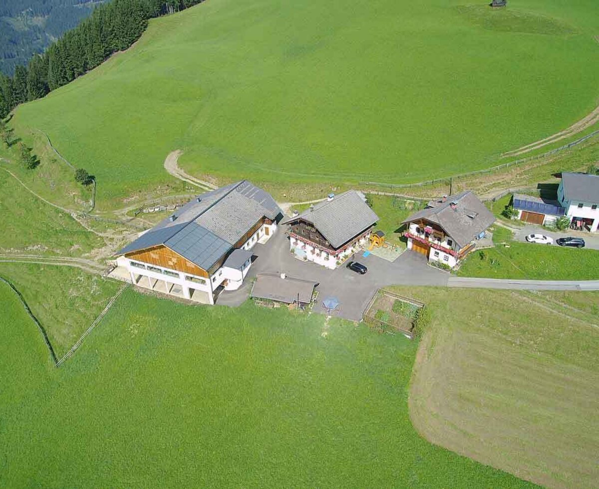 Luftaufnahme vom Bauernhof, mit mehreren Gebäuden, umgeben von grünen Wiesen und Bergen.