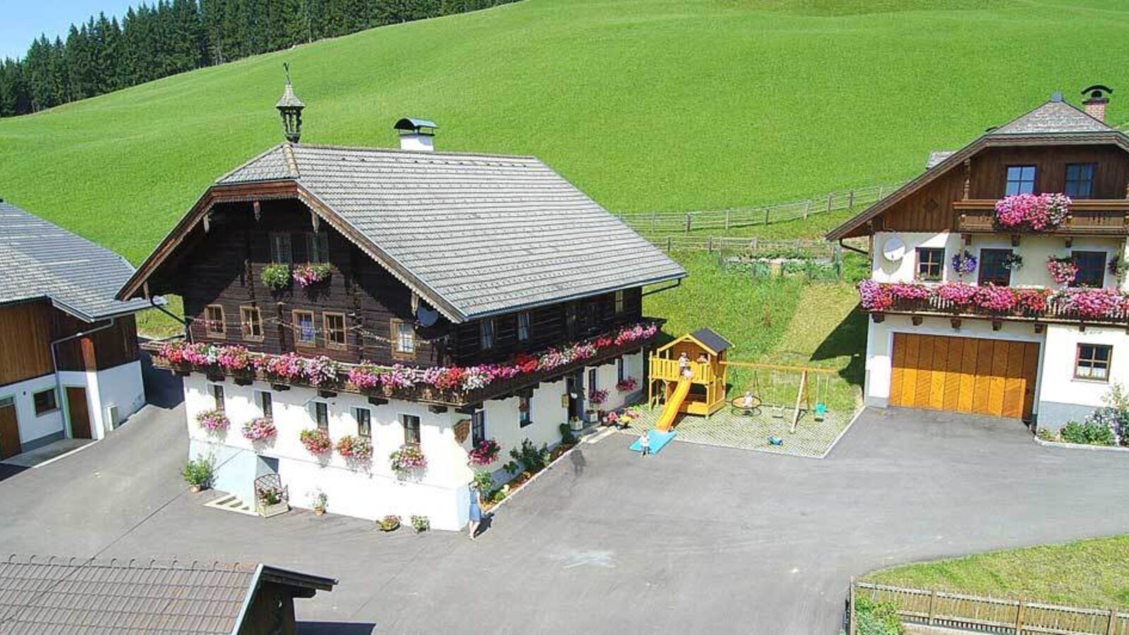 Der Bauernhof mit traditionellen Gebäuden, Blumenkästen, einem Kinderspielplatz und grünen Hügeln im Hintergrund.