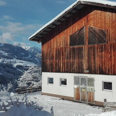 Das Gebäude des Bauernhofs mit seiner Holzfassade im oberen Teil und dem Ausblick auf die verschneite Berglandschaft.