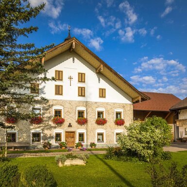 Die Außenansicht des Bauernhofs mit traditioneller Architektur, Fenstern mit Blumenkästen und grünem Gartenbereich.