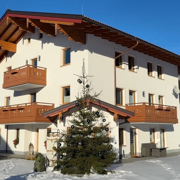 Außenansicht des Bauernhofs mit mehreren Balkonen, einem geschmückten Weihnachtsbaum und schneebedeckter Berglandschaft.