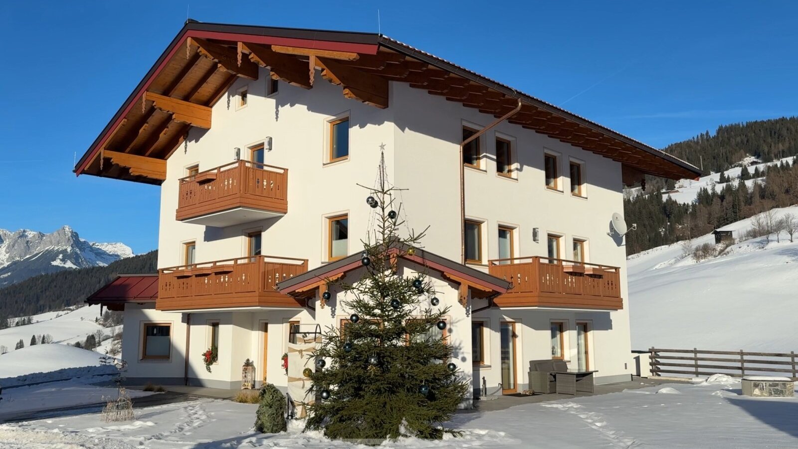 Außenansicht des Bauernhofs mit mehreren Balkonen, einem geschmückten Weihnachtsbaum und schneebedeckter Berglandschaft.